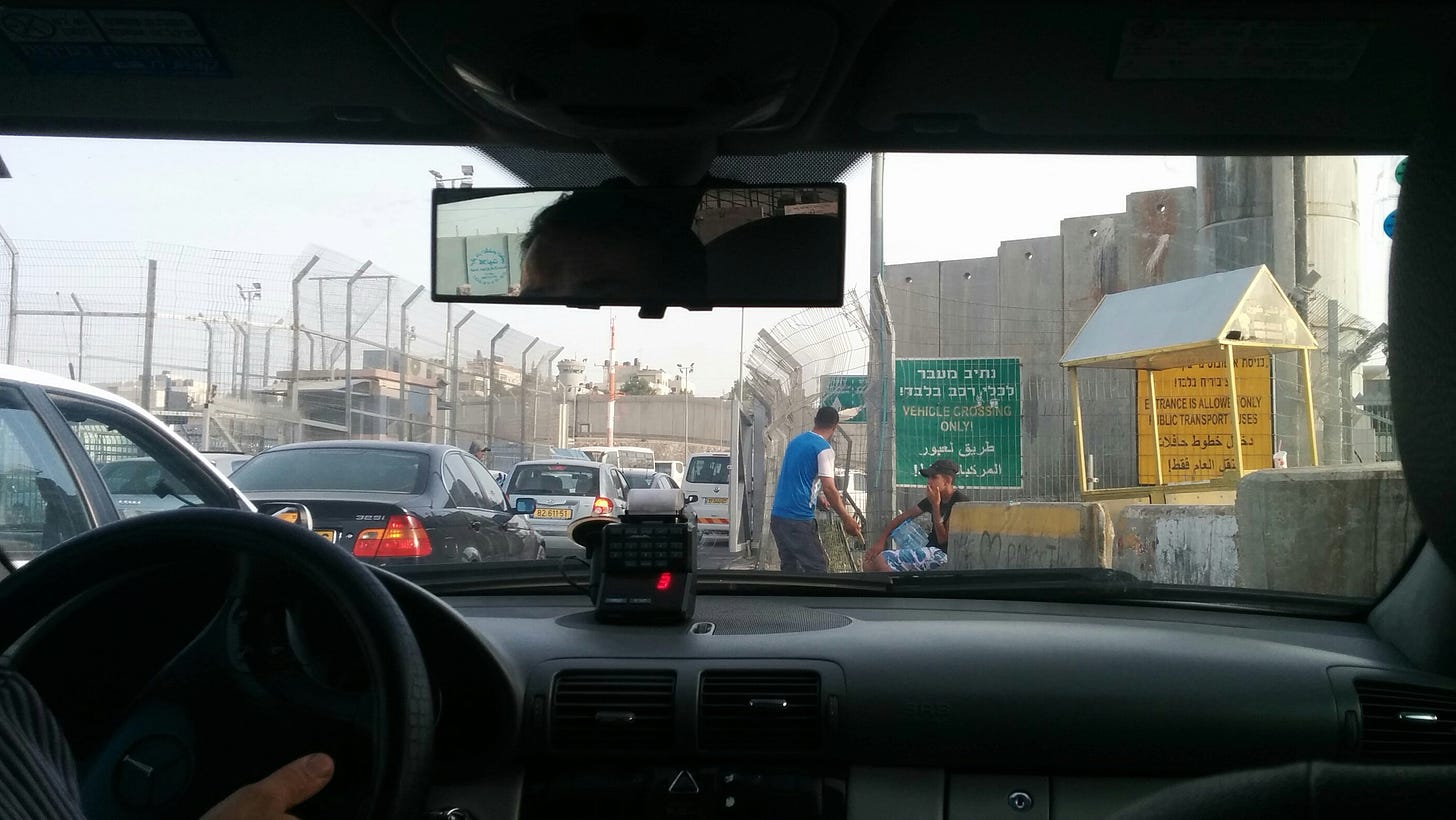 View out a car windshield of a border crossing lined with tall fencing and concrete walls, and two men waiting on the side of the road