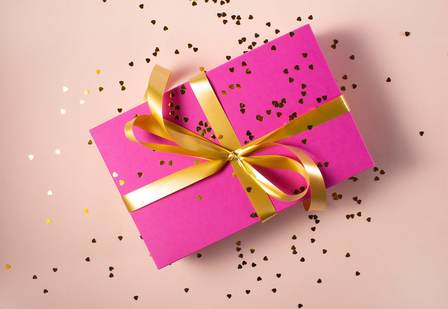 A bright pink gift box with gold ribbon and sprinkled with gold heart confetti