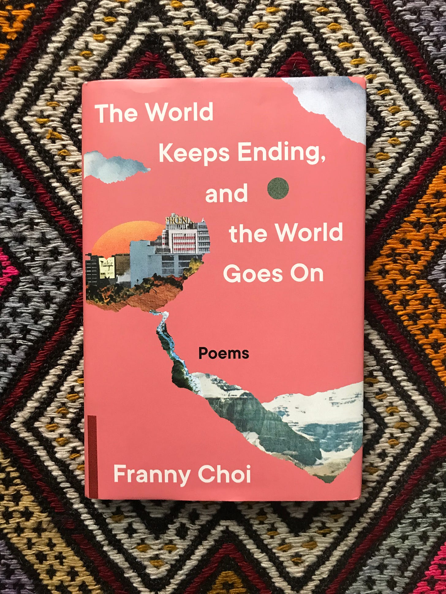 Picture of a book sitting on a rug. The book is called "The World Keeps Ending, and the World Goes On: Poems" and the cover is mostly pink, with an image of buildings on a mountain with the sun and a cloud behind them.