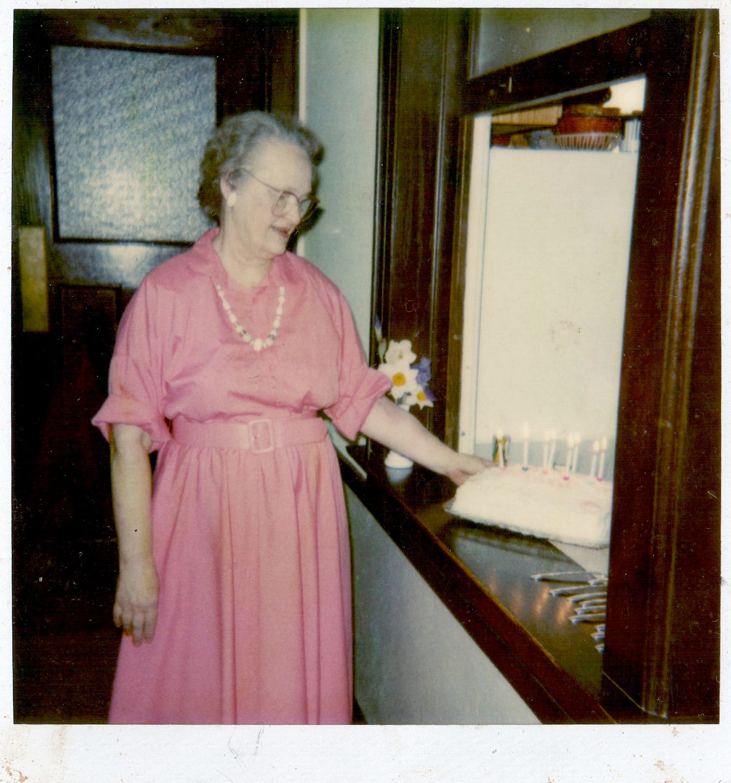 A polaroid photo of Rebekah's grandma next to a cake with lit candles. She is wearing a long pink dress with a belt and rolled up sleeves, a white beaded necklace, and her iconic clip on earrings. Her hair is short and gray. She's looking at her cake. This was one of her last birthdays. A polaroid photo of Rebekah's grandma next to a cake with lit candles. She is wearing a long pink dress with a belt and rolled up sleeves, a white beaded necklace, and her iconic clip on earrings. Her hair is short and gray. She's looking at her cake. This was one of her last birthdays.