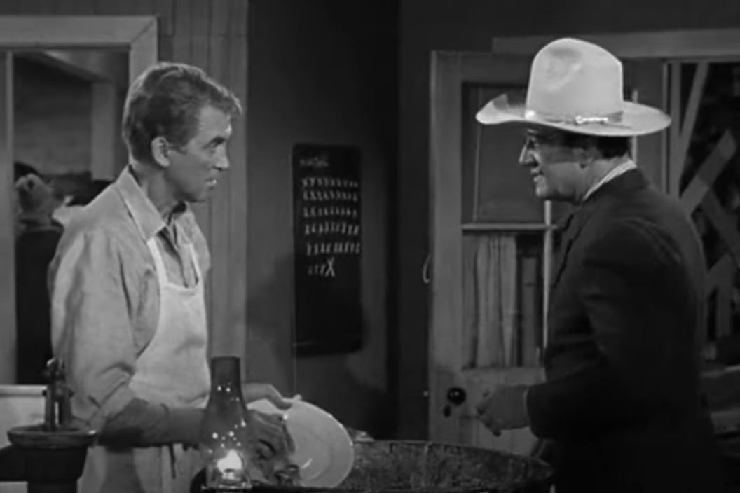 Still from The Man Who Shot Liberty Valance. Jimmy Stewart washes dishes in an apron while John Wayne talks at him.