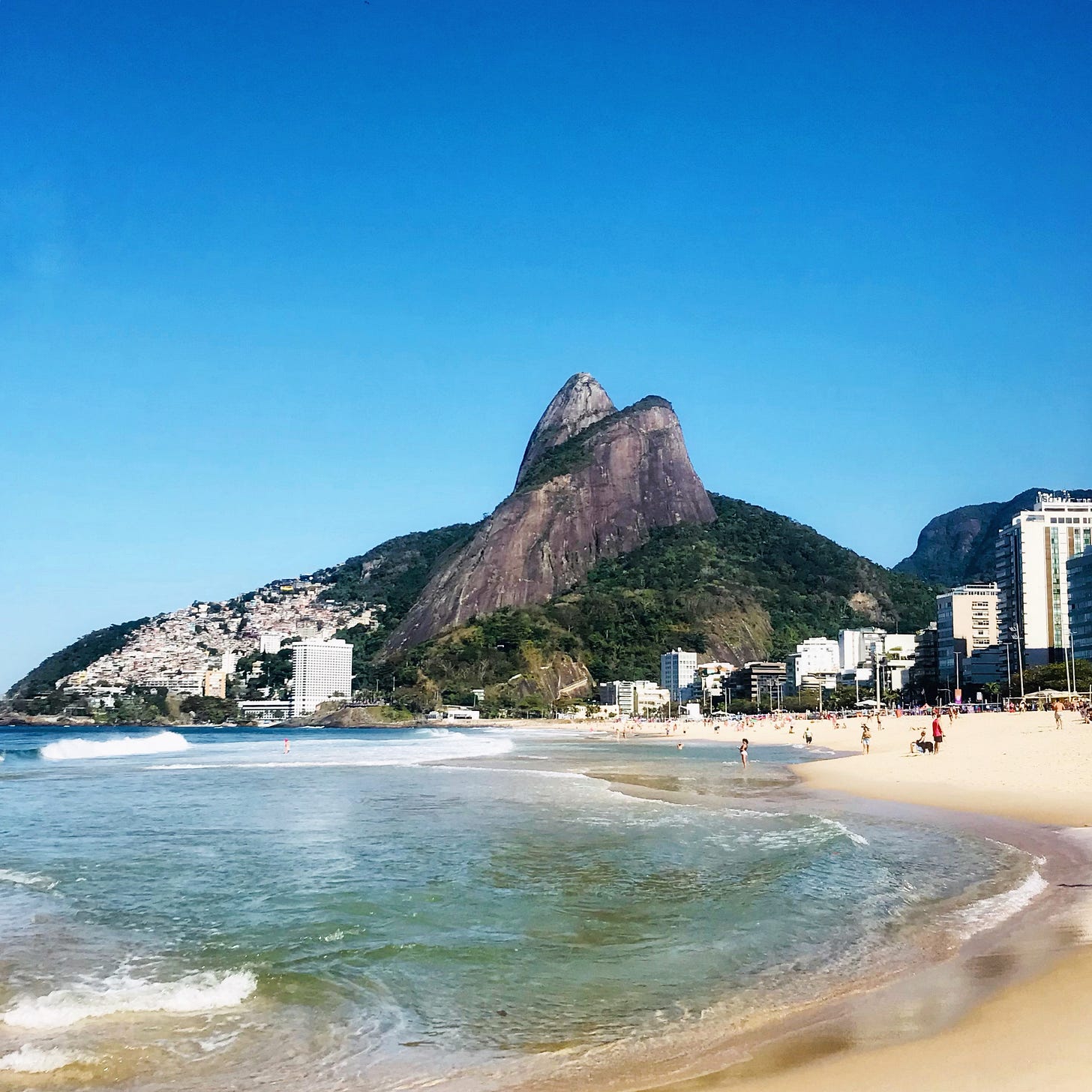 Picture of a beach, in Rio, with blue skies, a hill on the horizon, the sea to the left, and sand and some buildings to the right.