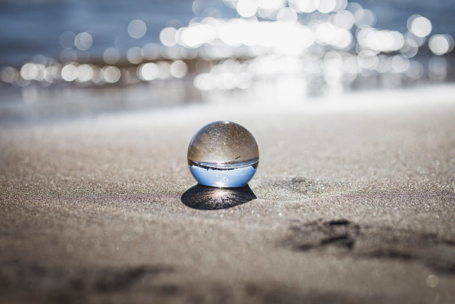 Piccola bocca di vetro appoggiata su una spiaggia, con lo sfondo del mare sfuocato e luminoso. Dentro alla sfera si riflette il paesaggio retrostante della costa.