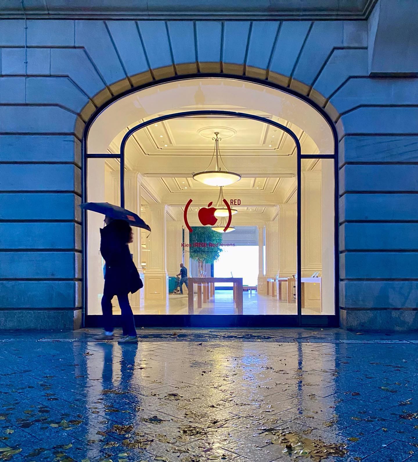 A person with an umbrella walks past the windows at Apple Amsterdam on a soggy day.