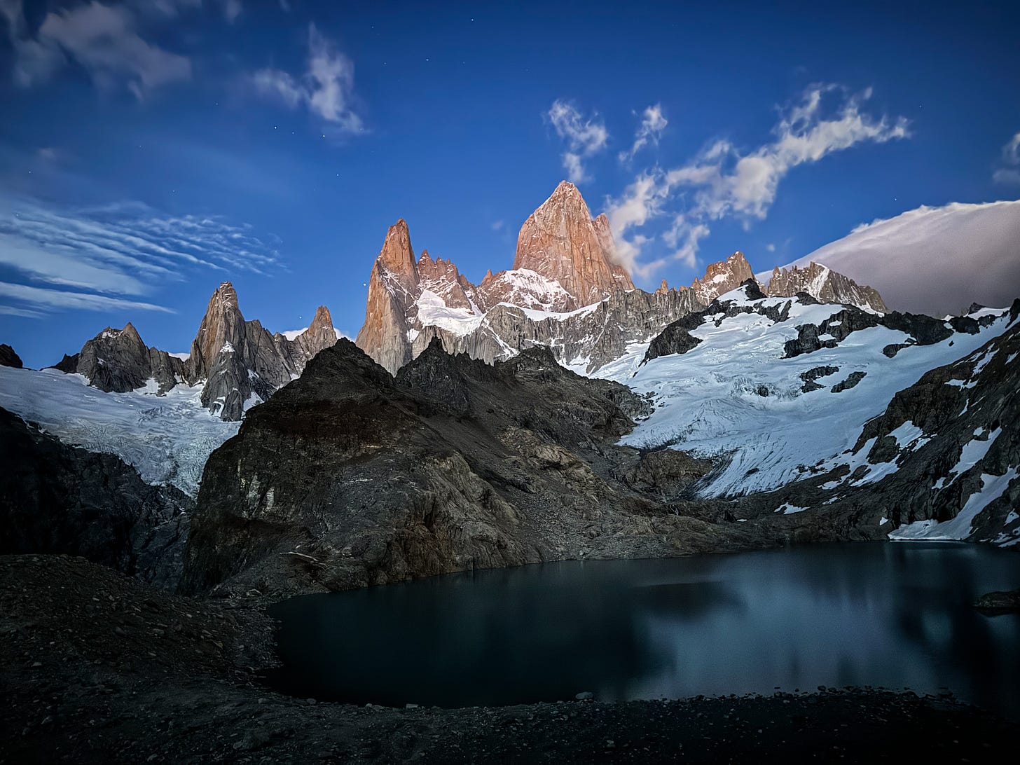 A line of granite towers rises up against a lightening sky. A dark lake is in the foreground, surrounded by white glaciers and smaller granite mountains. There are stars visible in the sky, with some clouds.
