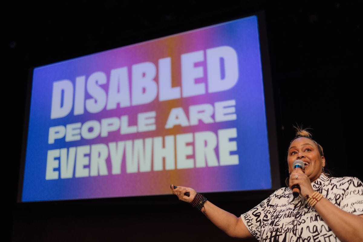 An Afro-Latina femme is speaking into a mic in front of a slide that says Disabled people are everywhere.