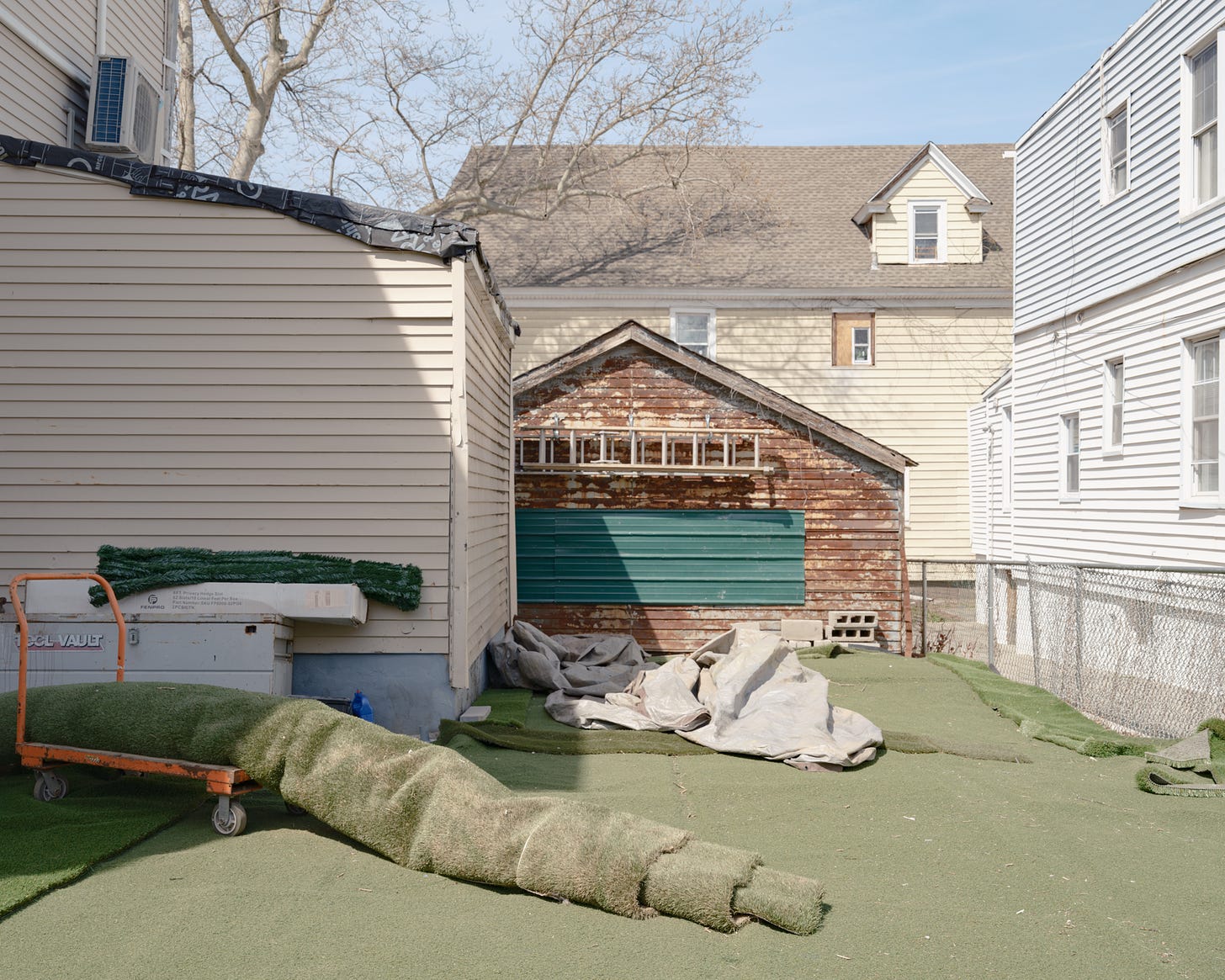 Large roll of green astroturf lies on top of orange cart in backyard of Woodside homes