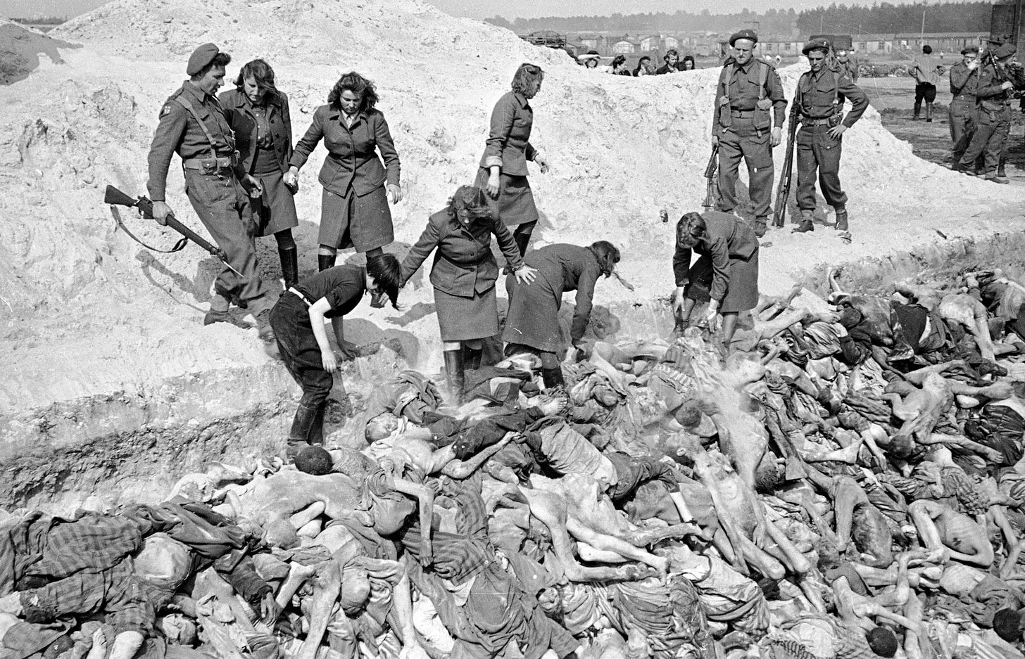 Black and white photo of a pit of sketetal corpses, Nazis hovering and picking though the bodies.