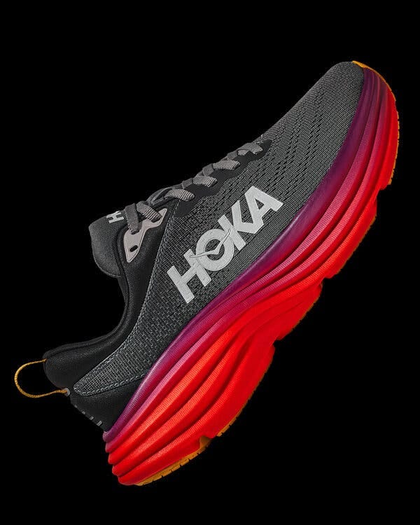 A black and gray running shoe with a bight red and purple sole, at a diagonal with the toe pointing up toward the upper right corner of the frame on a black background.