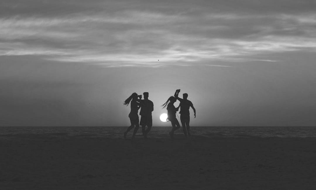 silhouette photo of four people dancing on sands near shoreline silhouette photo of four people dancing on sands near shoreline