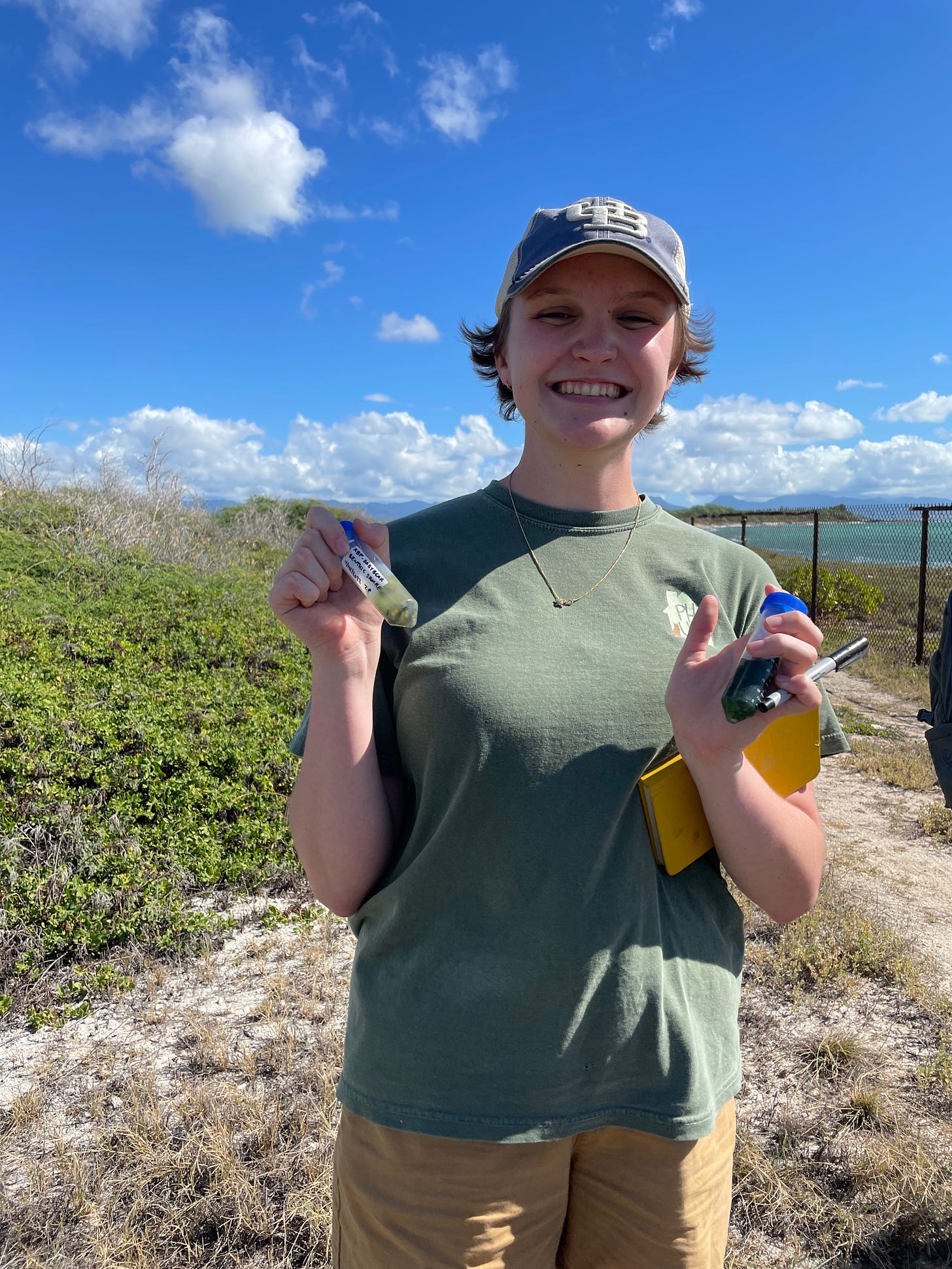 Image Description: Zoe, a white nonbinary person, is beaming towards the camera and holding far too many things ( including a notebook, a pen, and two tubes of green algae) on the coast of Oahu. The ocean is visible in the background with ground cover plants and a dirt road closer to the foreground.