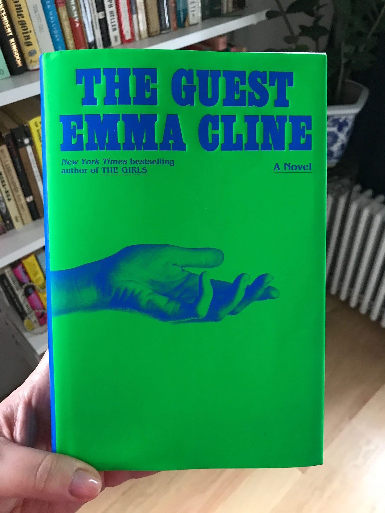 Holding a copy of Emma Cline's The Guest, which is bright green and blue and has the image of an outstretched hand on the cover.