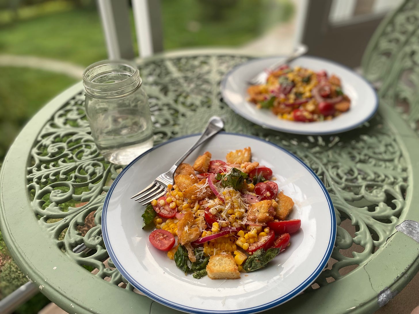 On a green café table, two plates of the panzanella described above, on a white plate with a blue rim: croutons, red onion, grape tomatoes, basil, and yellow corn, topped with pecorino and black pepper.