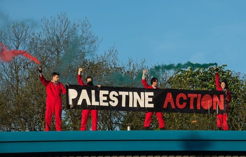 Four people in boiler suits on a rooftop holding a "Palestine Action" sign