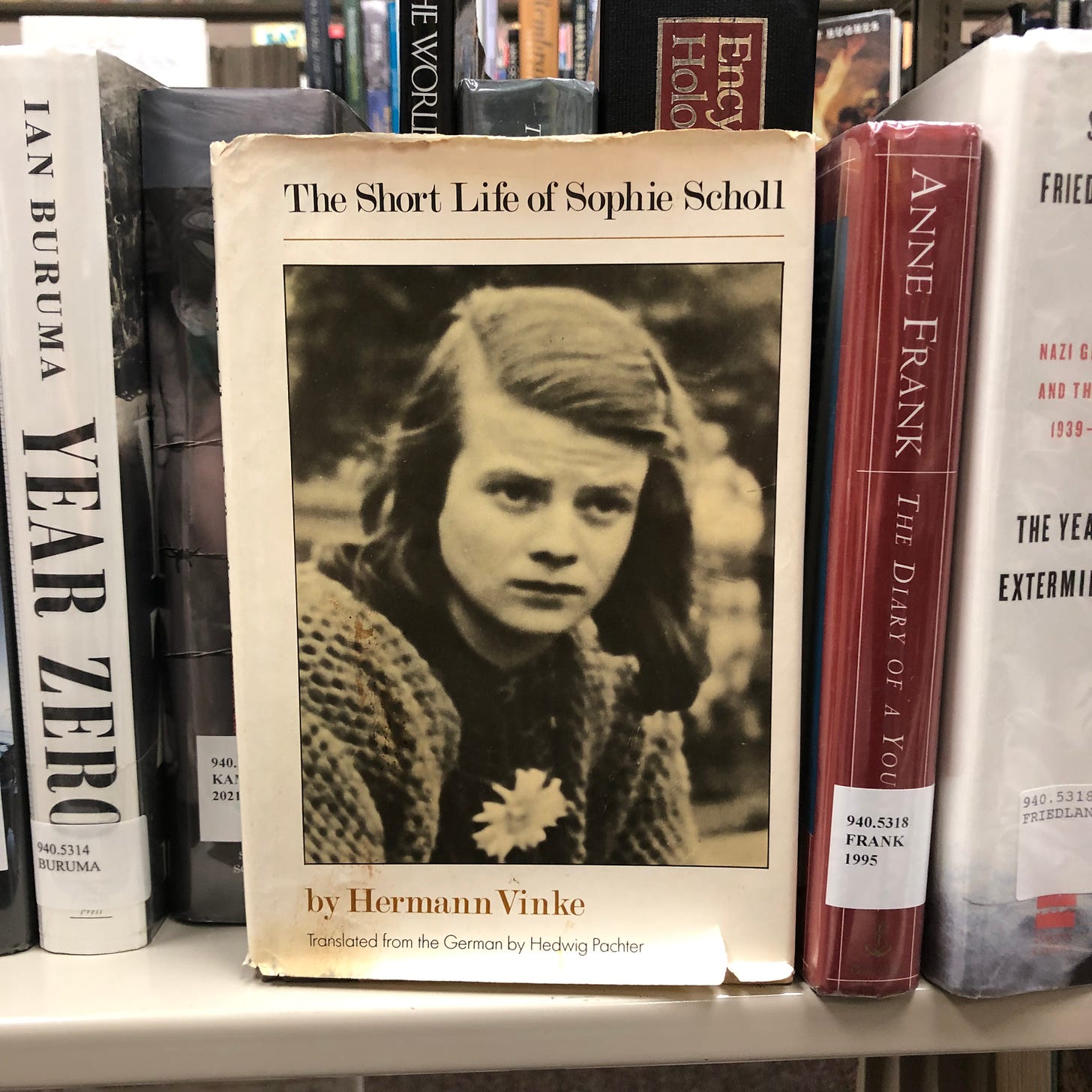 Picture of book on library shelf -- The Short Life of Sophie Scholl by Hermann Vinke, showing the iconic b&w picture of Sophie staring directly out on the front cover.
