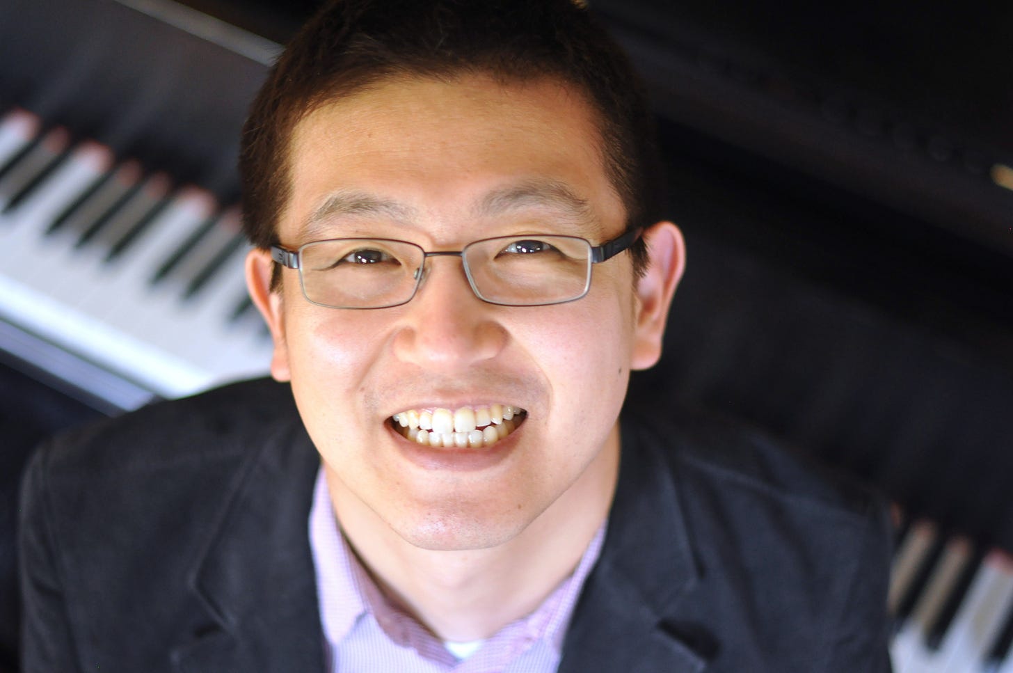 Keigo Hirakawa, a Japanese-American man, wearing a dark jacket, purple shirt, and wire-rimmed glasses, seated at a piano, and smiling up at the camera