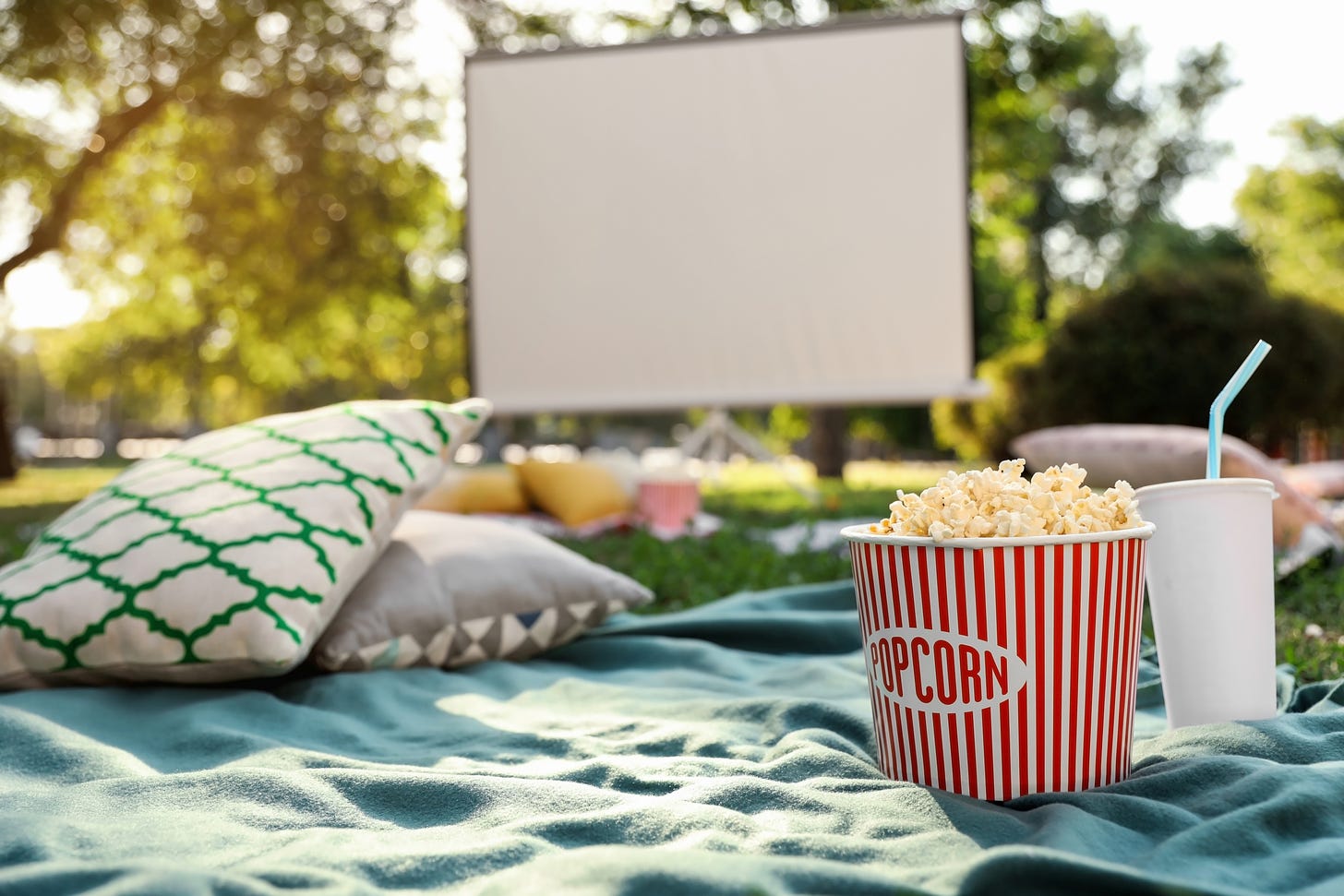 Low view point of an open air cinema. Blanket over the grass, and on it there are two cushions, a red-and-white bucket of popcorn, and a white soda cup. In the background there’s a big white movie screen.