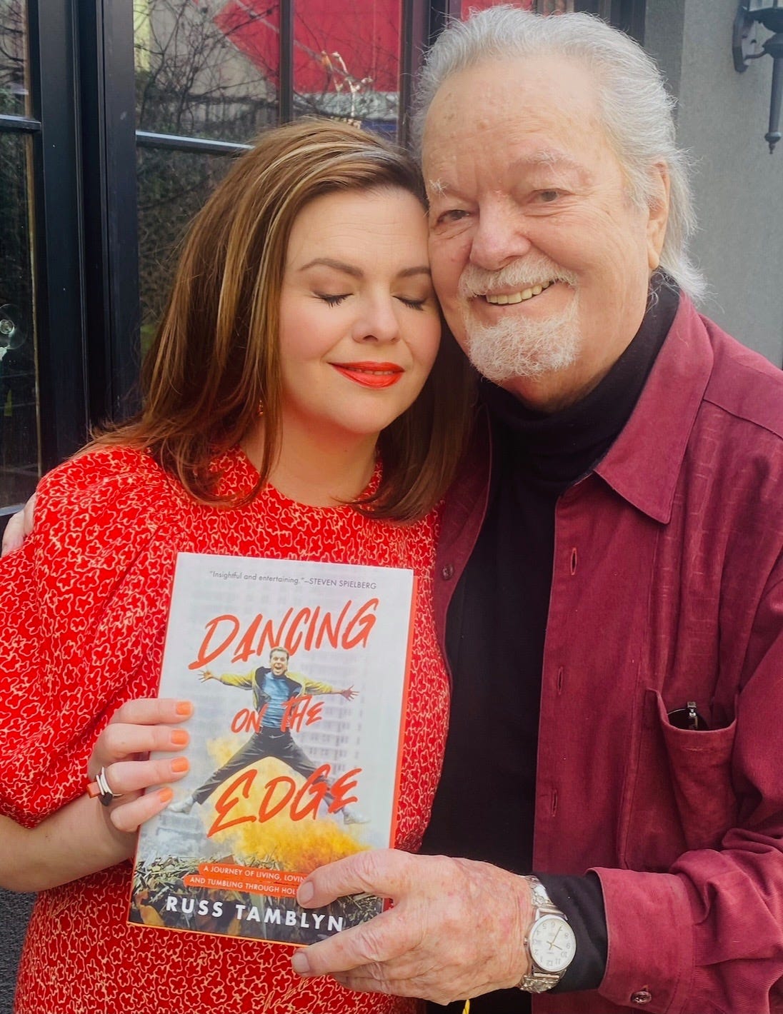 Amber and Russ Tamblyn hold Russ' memoir, "Dancing on the Edge." Russ smiles into the camera. Amber has her eyes closed and a soft smile with the head pressed against her dad's cheek.