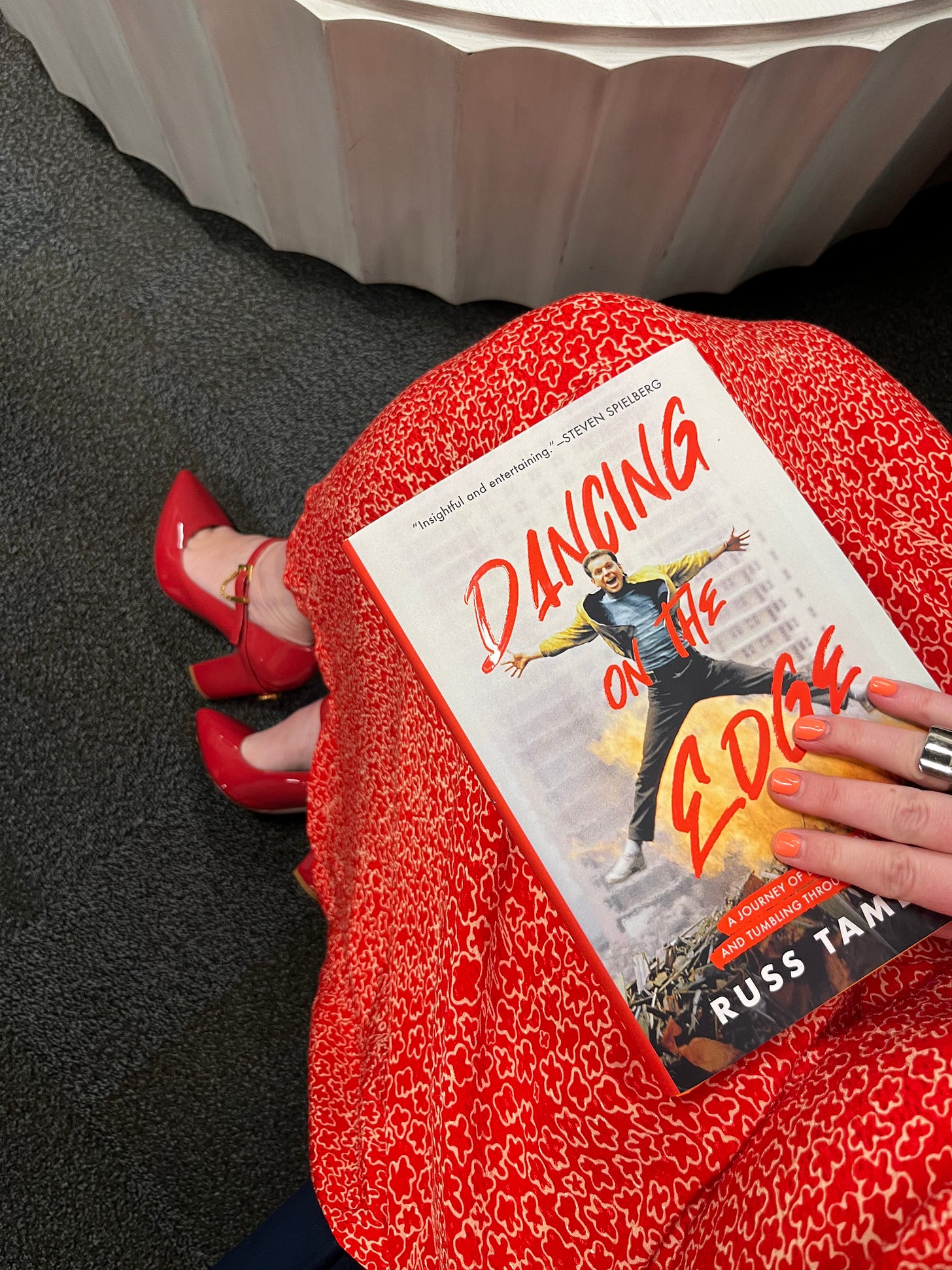 A photo of the book resting on Amber's lap. She wears a red-orange dress with matching shoes and orange nail polish which all color coordinate with the font color on the book cover.