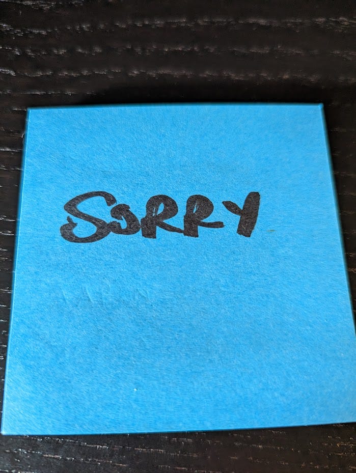 A blue sticky note on a black desk, the word "sorry" in black ink