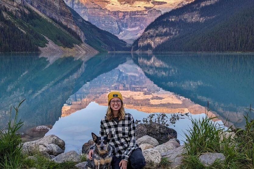 Haley, a young woman in a yellow hat, and Scout, a blue heeler wearing a white leash, sit on the shore of Lake Louise in Banff National Park