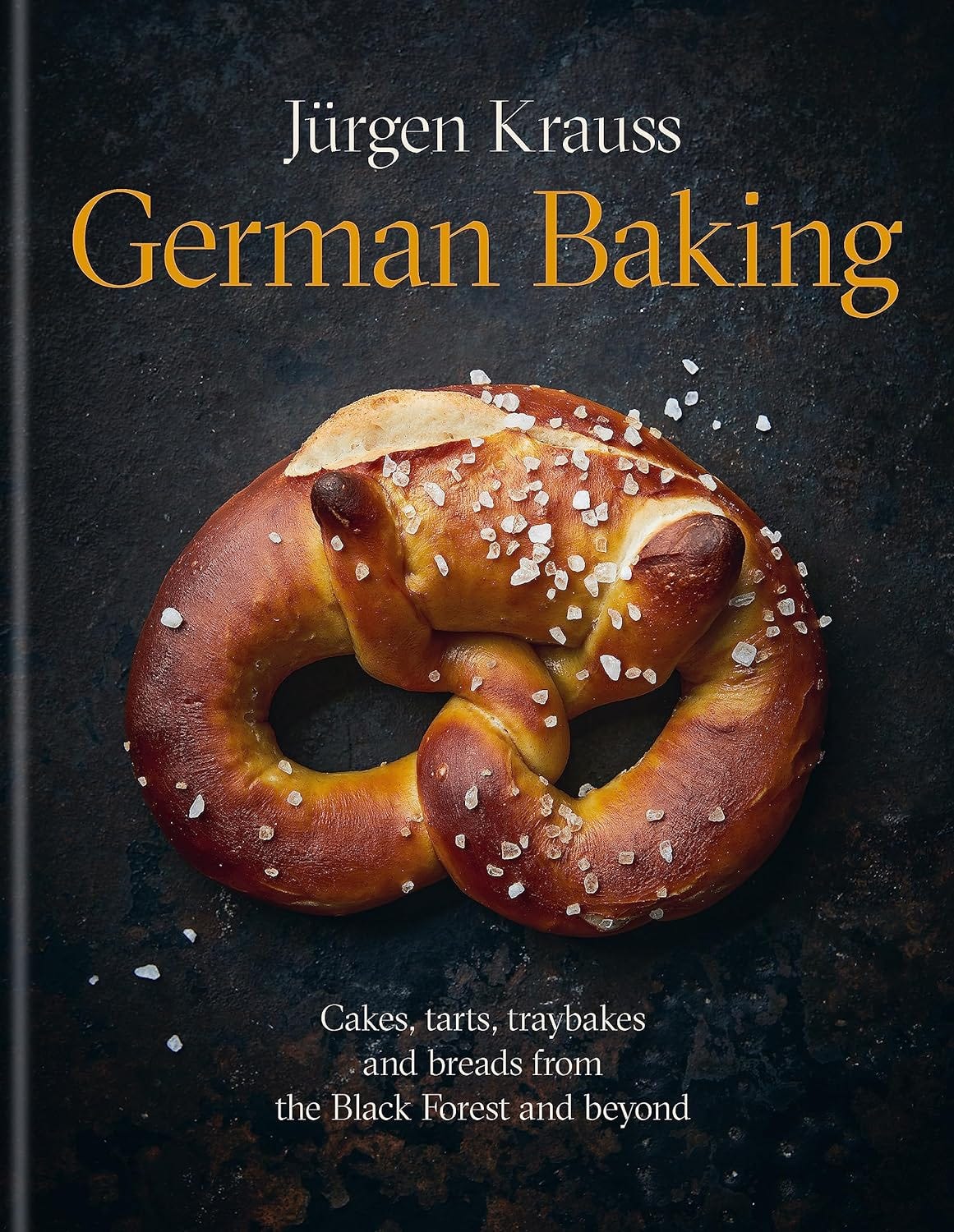 The cover of German Baking. Gold and white text on a black background with a close-up of a  rocksalt-encrusted twisted pretzel in all its burnished glory