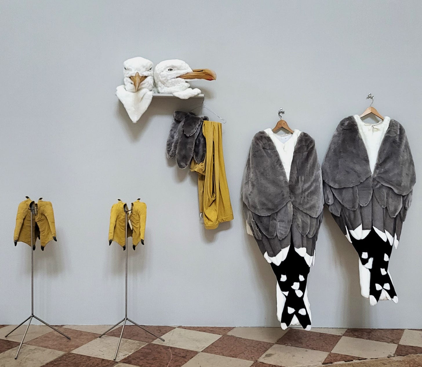 Set against a grey wall, two seagull costumes hang on racks. The two heads of the costumes are displayed on the shelf.