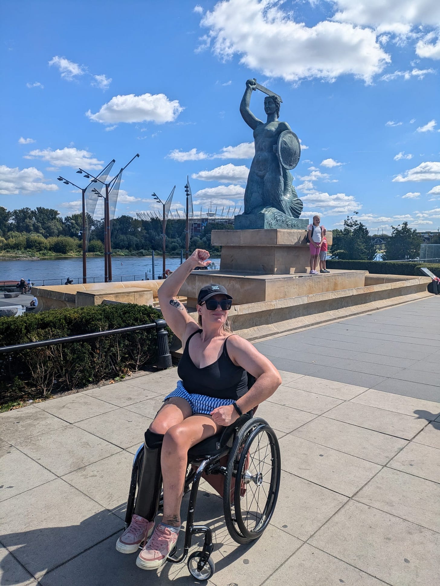 It's a sunny day with blue skies and white clouds, I pose in front of the Mermaid of Warsaw, a stone statue that looks over the River Vistula. I mimic her pose with one arm in the air holding a sword, and the other posing as if holding a shield. 