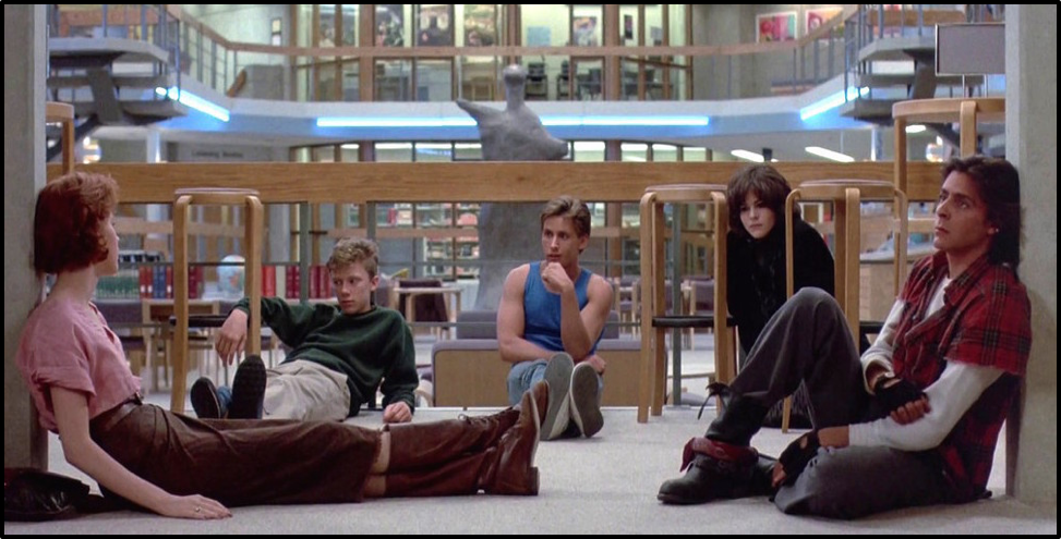 Still from The Breakfast Club. The five high school students sit on the floor of the library, not working on their essay for detention.