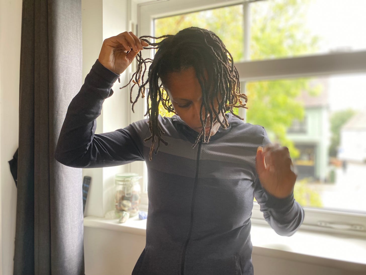 A Black femme with locs twists her locs, head down standing in front of a window wearing grey long sleeved shirt