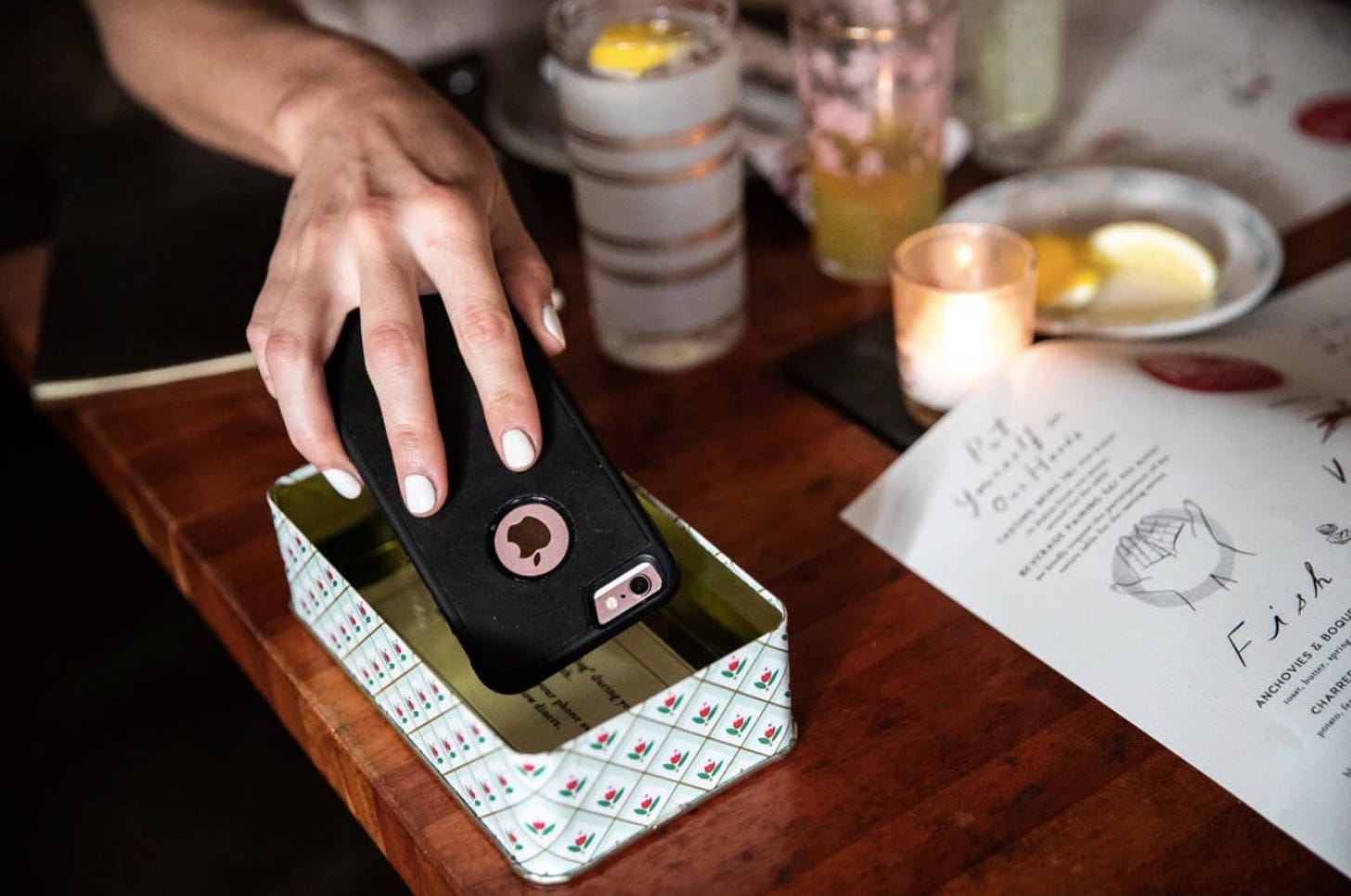 A woman's hand places a phone face-down into a tin antique box on a restaurant table.