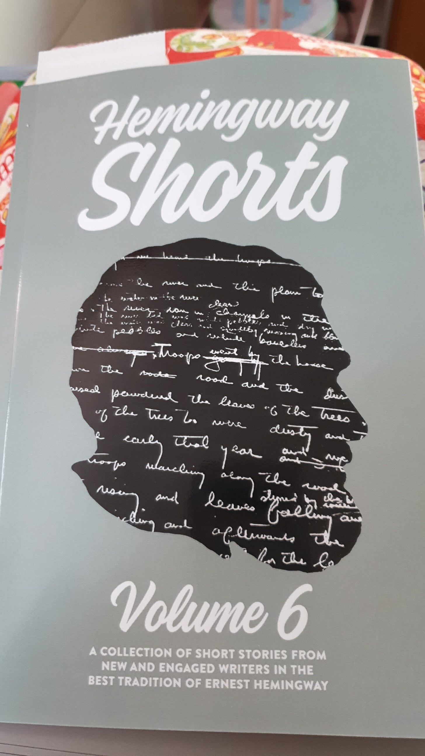 Image shows the book cover, a grey background with the title ‘Hemingway Shorts’ at the top then a black silhouette face shape of Hemingway with his handwriting in white lettering. At the bottom it reads ‘Volume Six a collection of short stories from new and engaged authors in the best tradition of Ernest Hemingway’.