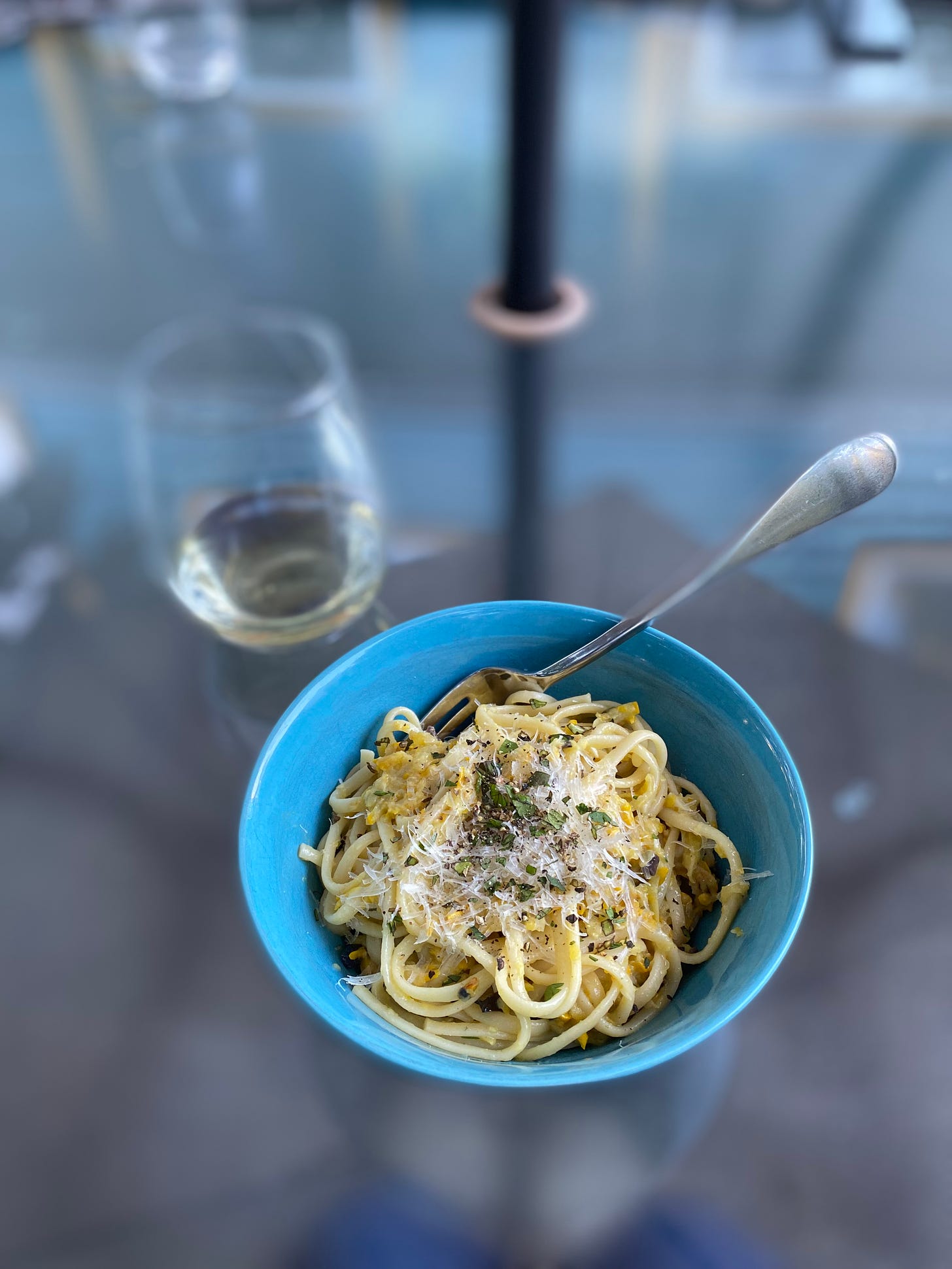 A blue bowl of linguine in zucchini butter sauce. On top are chopped herbs and grated parmesan, and a fork rests at the back of the bowl. Behind it and to the left is a stemless glass of white wine.