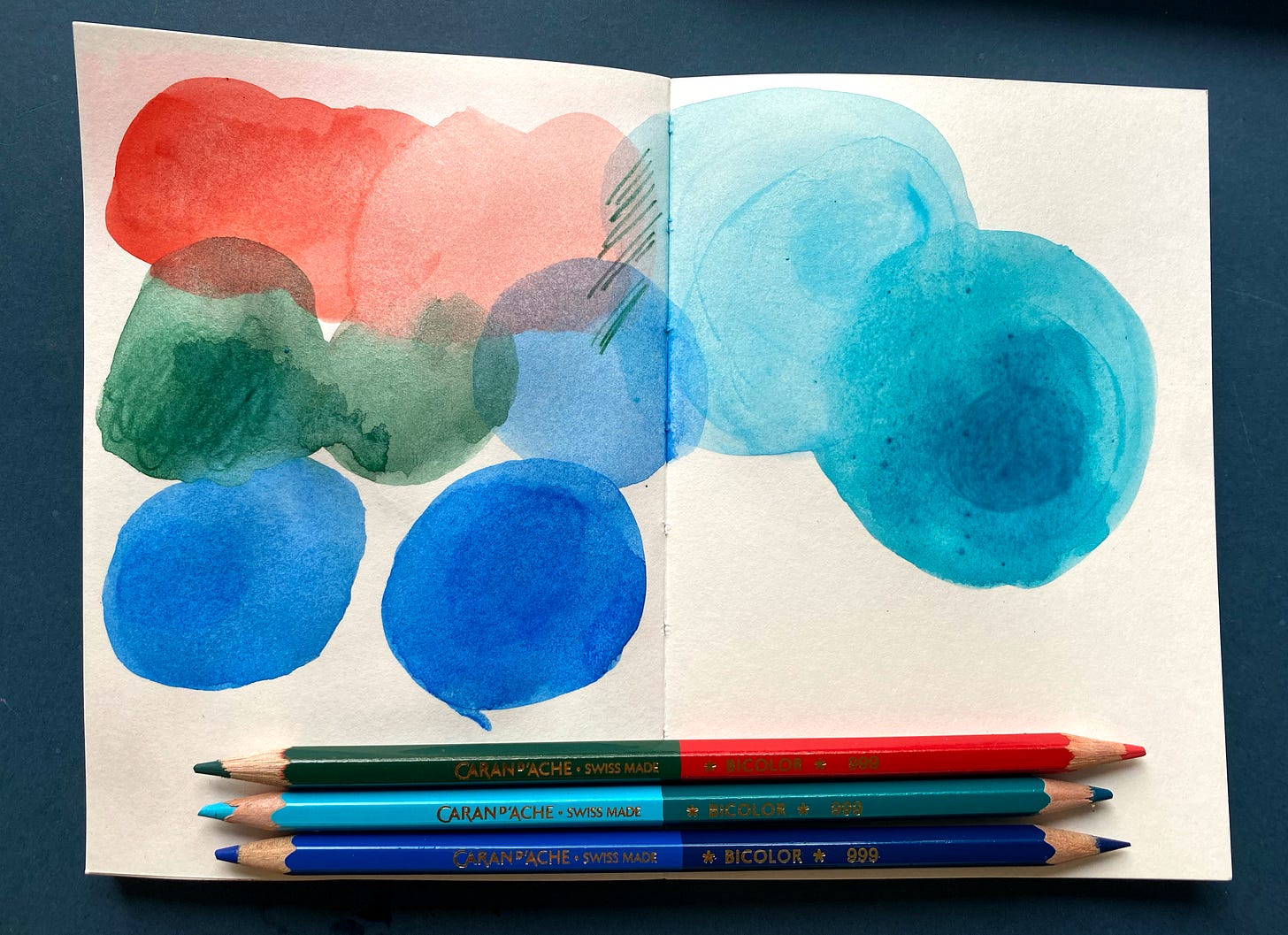 sketchbook page open showing colour swatches of tones of blue, green and vermillion. Water soluble pencils shown next to the page.