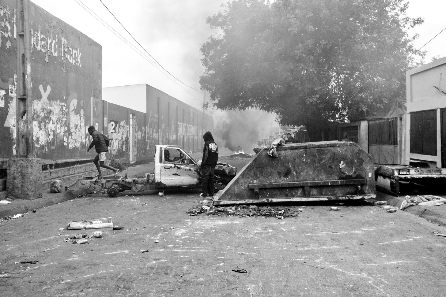 a man stepping on a destroyed truck near a trash bin in the middle of the street, there's another man wearing a jacket standing to the side