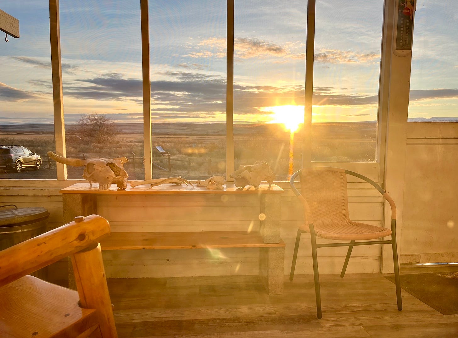 A sun rises over a distant horizon, as seen from a porch where skulls sit on a table next to a chair
