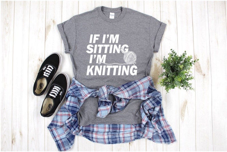 May include: A gray t-shirt with the text 'IF I'M SITTING I'M KNITTING' in white. The shirt is paired with a blue and red plaid shirt and black sneakers.