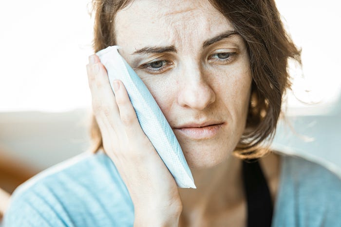 A woman who’s slightly miserable, holding up an ice-pack to her cheek in an effort to relief pain