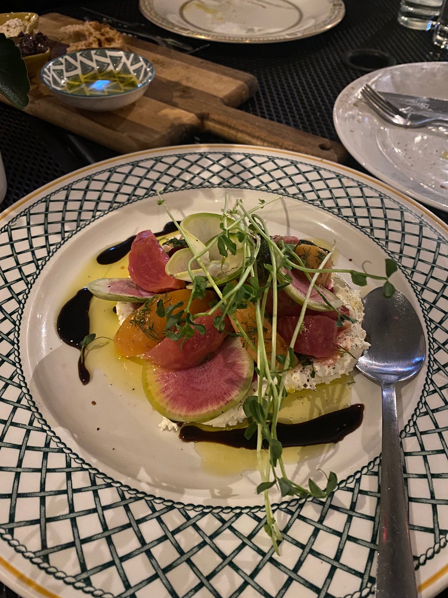 A white plate with a crosshatch patterned rim, with a brightly coloured beet salad on a bed of goat cheese, topped with pea shoots and ground pepper. There are puddles of oil and balsamic vinegar around the plate, and a large spoon resting on it. In the background is a bread board with a small piece of bread remaining.