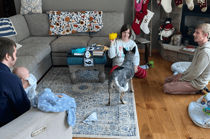 Scout the blue heeler plays fetch with a young woman in the living room of a house. Off to the side of the frame, a young baby watches from her father's lap