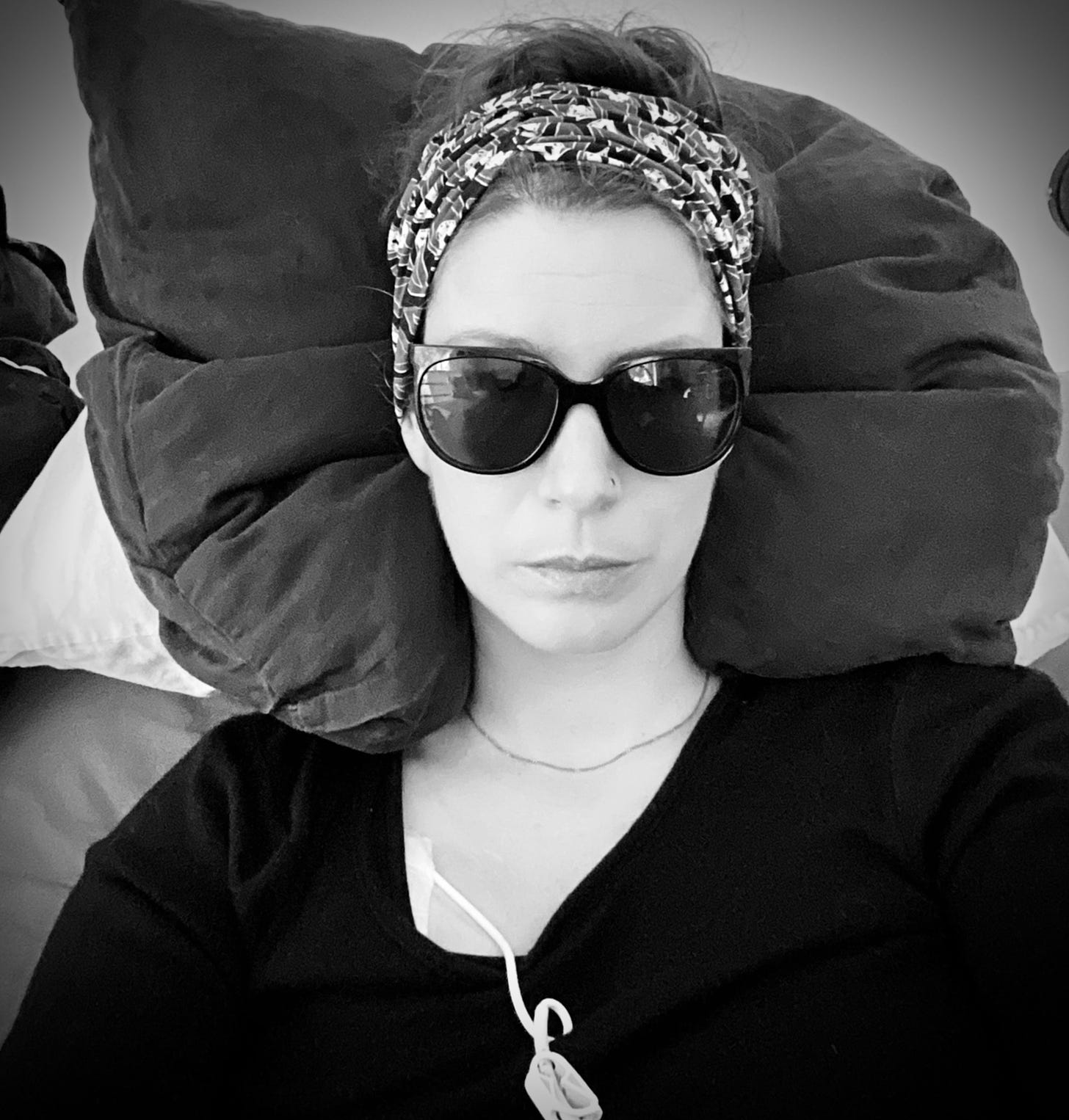 A white woman living with severe ME/CFS is lying in bed wearing sunglasses because of light sensitivity