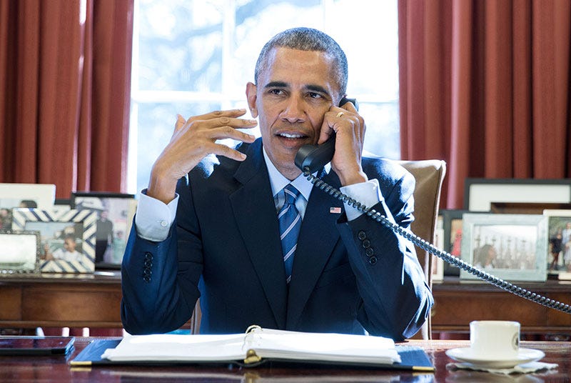 This image shows an undated photo of President Barack Obama sitting at the White House's Resolute Desk while talking on the phone with an unknown caller. Obama wears a blue suit, holds the phone receiver to his left ear, and gestures for emphasis with his right hand. This image shows an undated photo of President Barack Obama sitting at the White House's Resolute Desk while talking on the phone with an unknown caller. Obama wears a blue suit, holds the phone receiver to his left ear, and gestures for emphasis with his right hand.