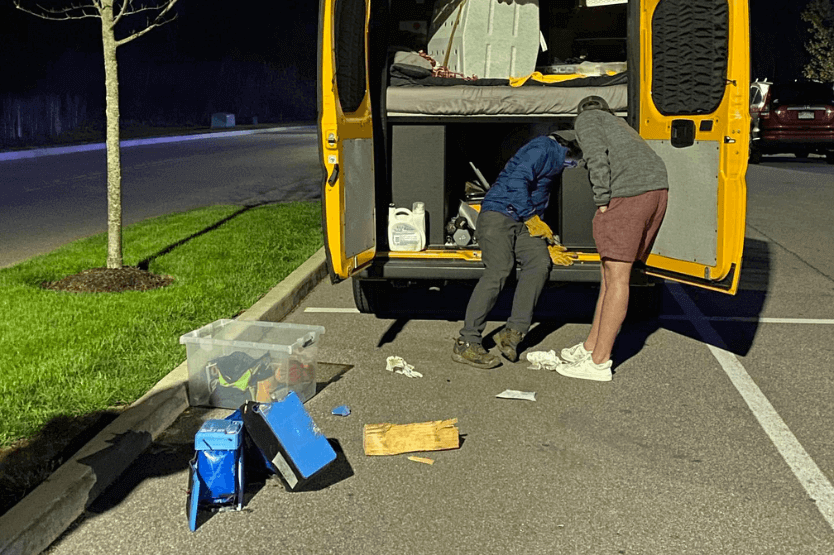 A yellow campervan sits in a parking lot at night with the rear doors open as two men stick their heads into the electrical cabinet inside. On the ground are two exploded lithium ion batteries that look a bit like accordions