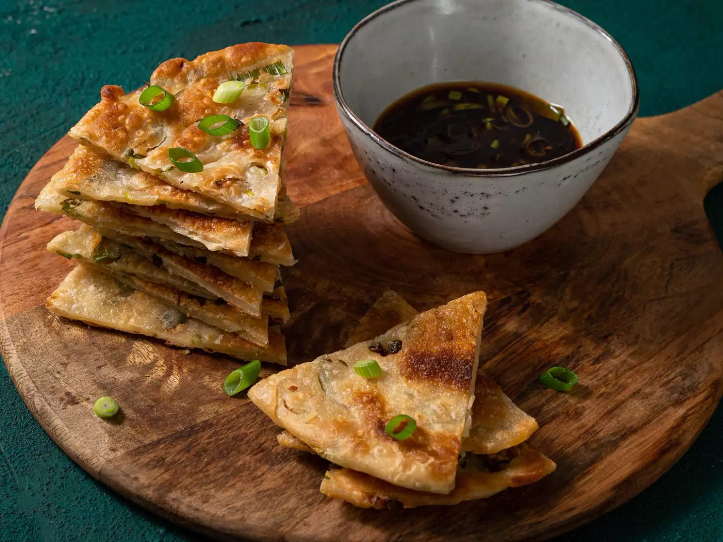 Scallion pancakes cut into slices, stacked on top of each other on a round wooden cutting board with a ceramic bowl of dipping sauce.