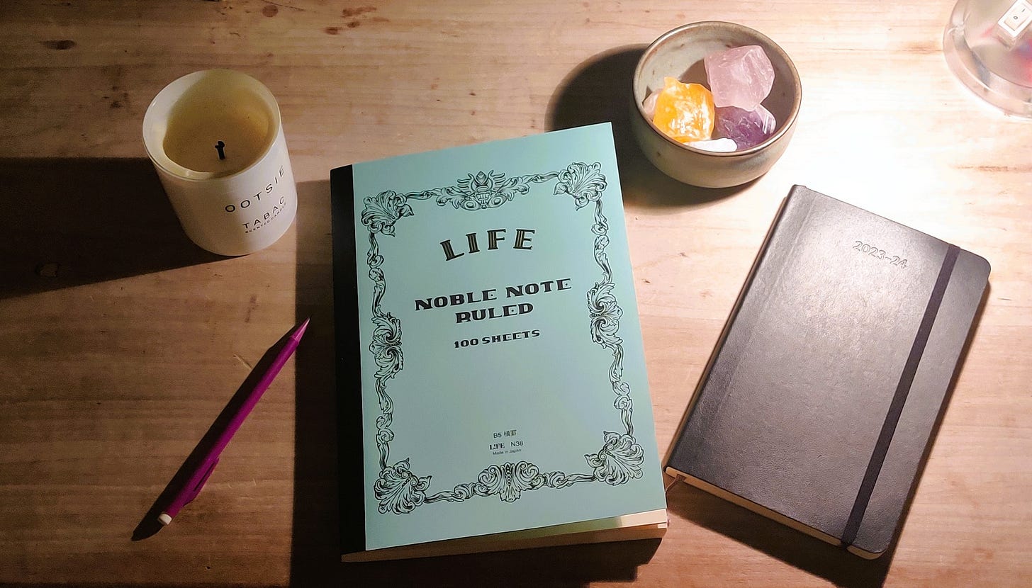 A wooden desk with a candle, a notebook and pencil, a bowl of crystals and a dairy on top.