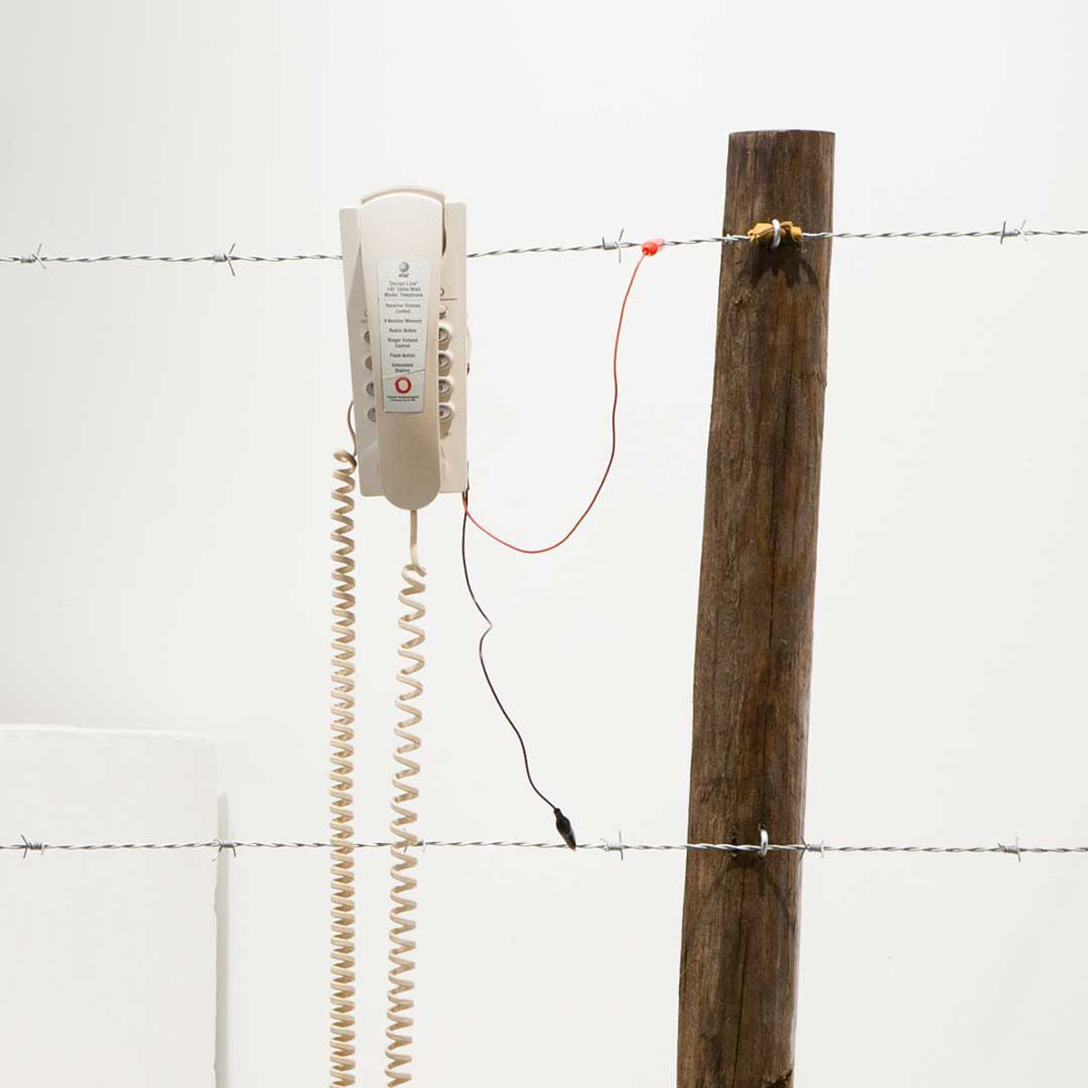 A fence post with two lines of barbed wire. A phone hangs from the top wire, and is connected to both with alligator clips.