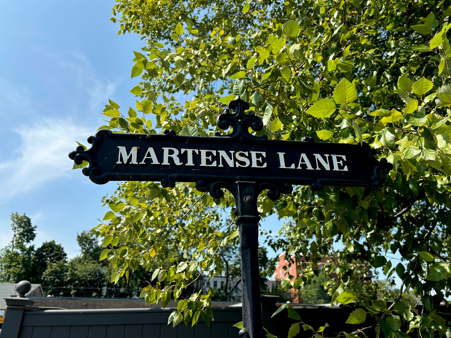 A decorate black sign with MARTENSE LANE written in white against green trees and blue sky.