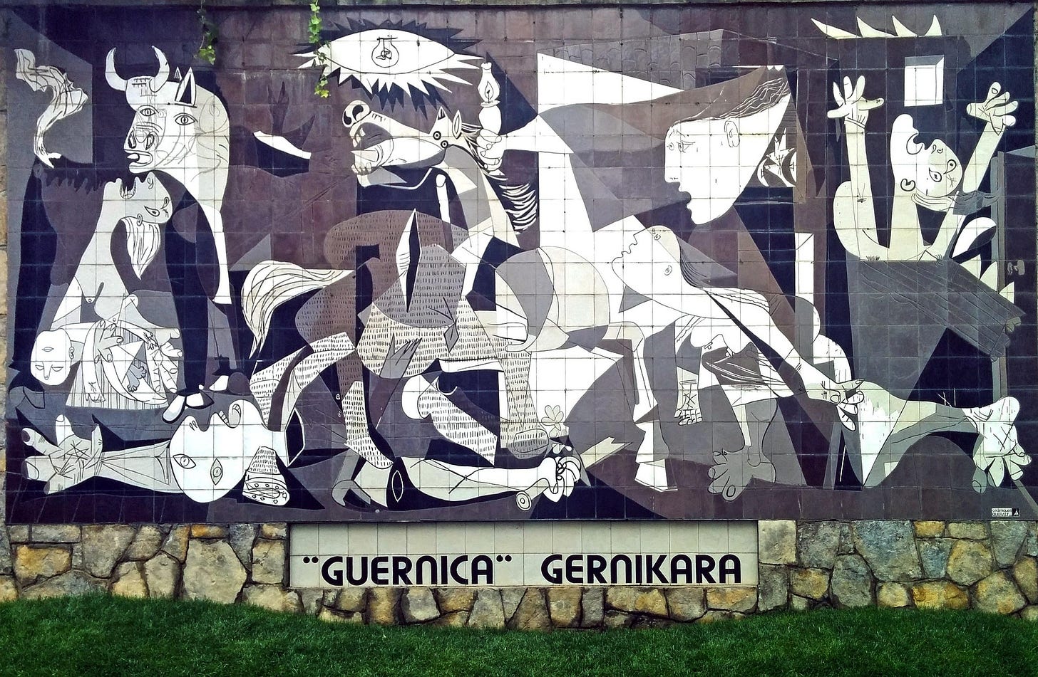 the full painting as a black and white mural. at the botom, above the green grass, the text "Guernica" Gernikara