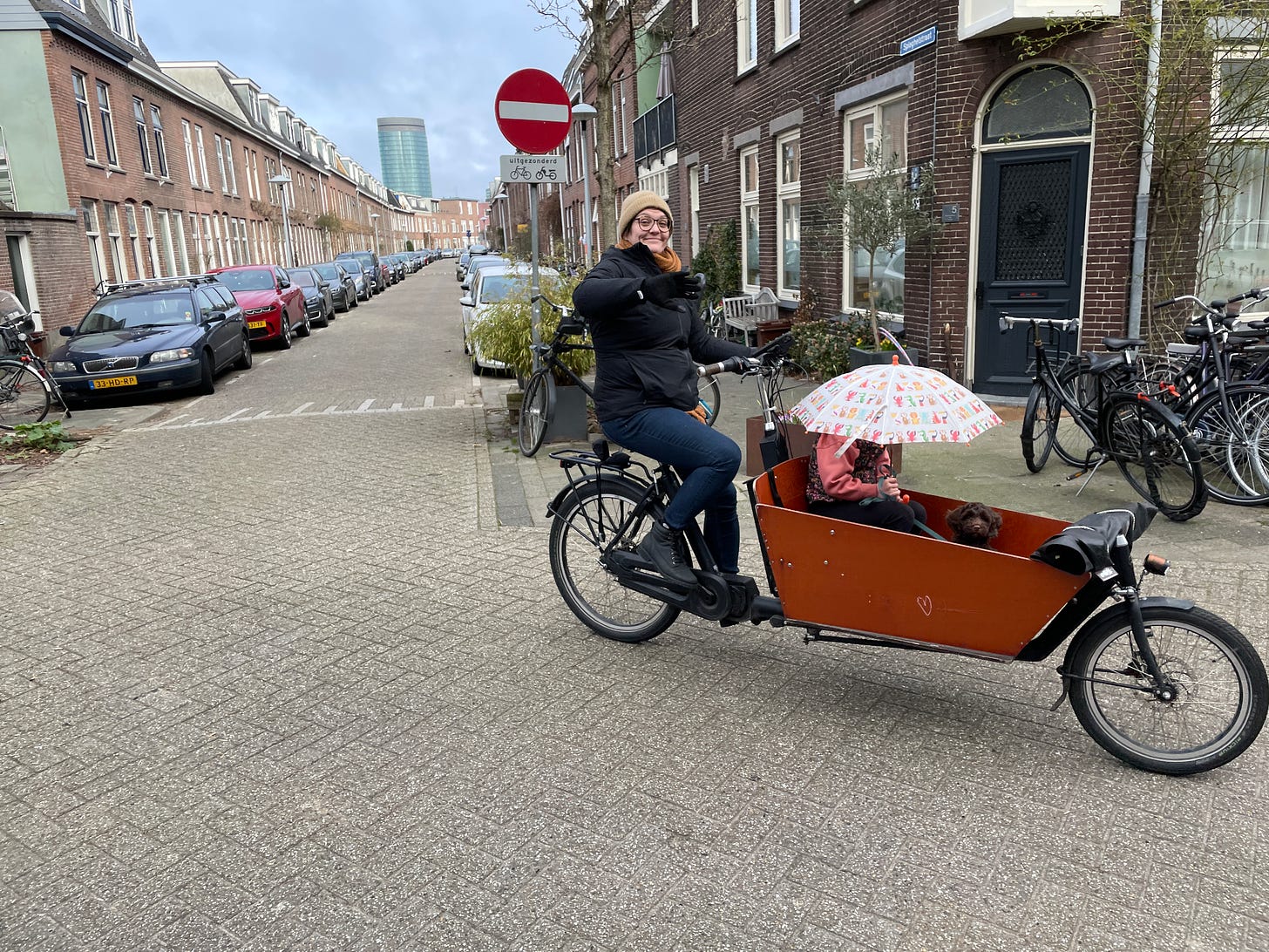 A woman rides a bakfiets cargobike down a dutch residential street. In the box are sat a young child and an adorable brown puppy