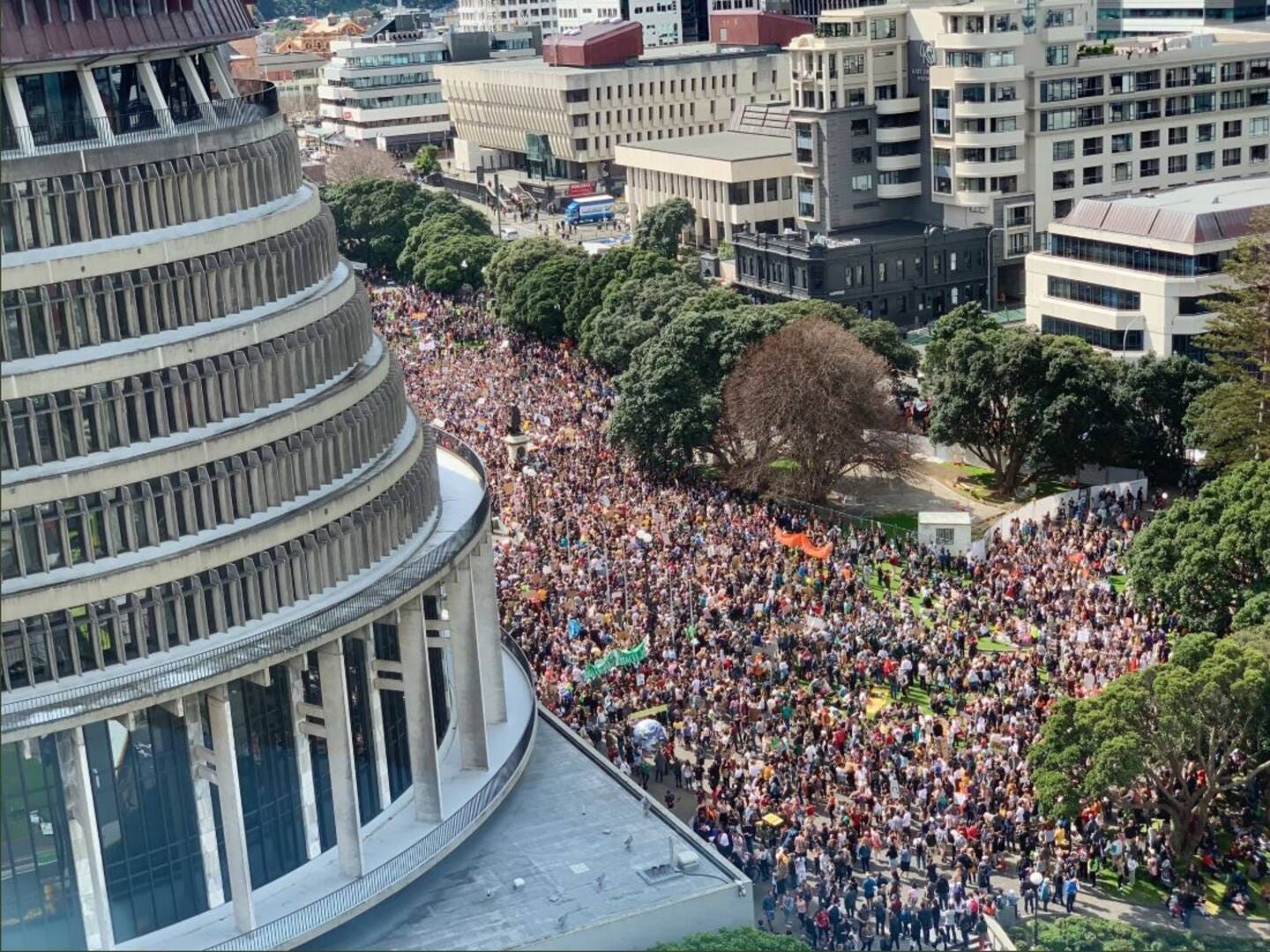 An aerial shot of the Parliament Lawn with the Beehive shown in the corner, packed with a crowd of people holding banners and signs.